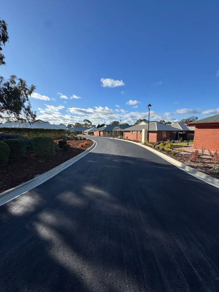 A freshly paved road curves through a residential neighborhood with red-brick houses, landscaped gardens, and a bright blue sky with scattered clouds overhead.